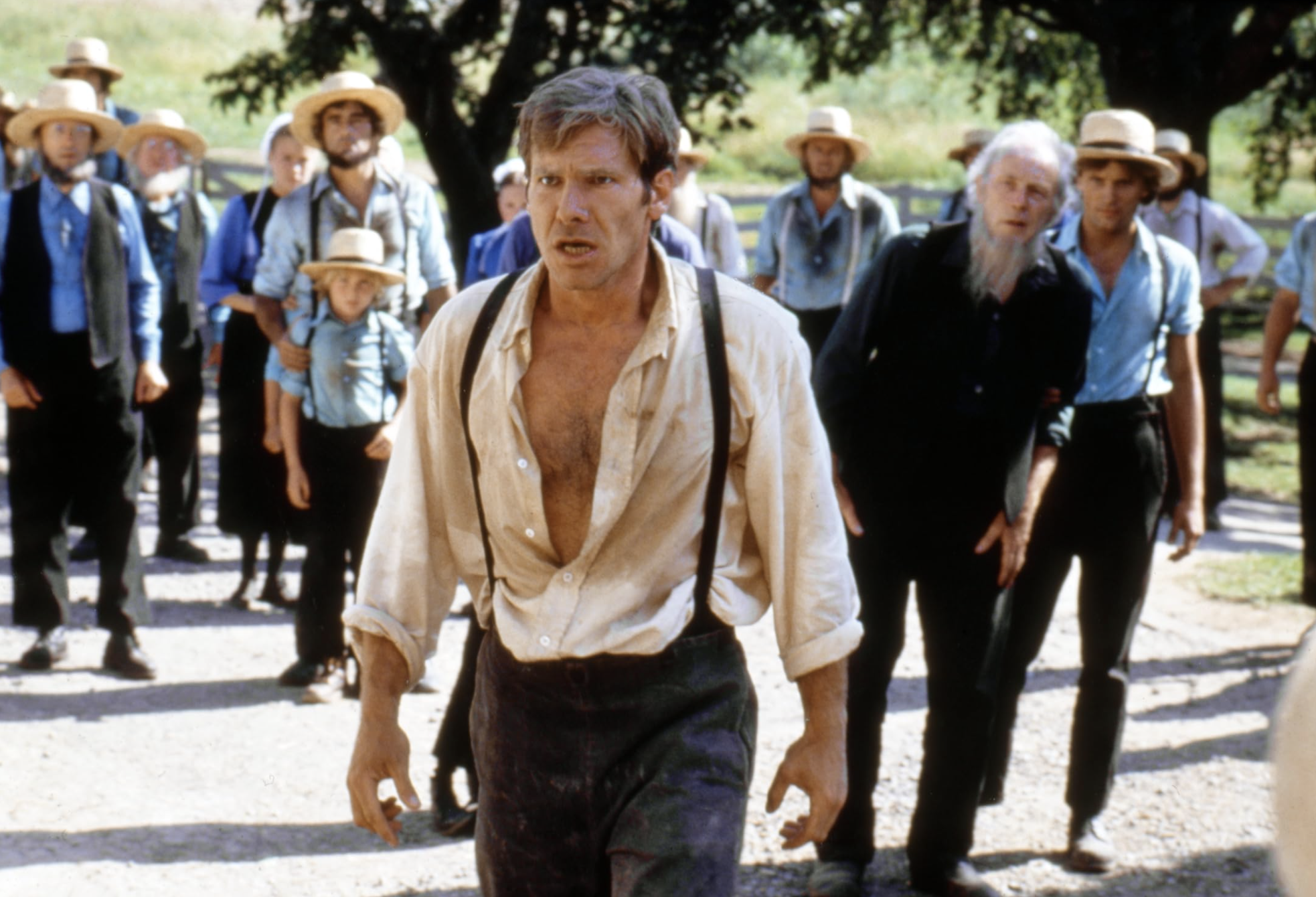 Harrison Ford in a ripped shirt, suspenders, and pants standing in front of a bunch of dudes in the movie WITNESS